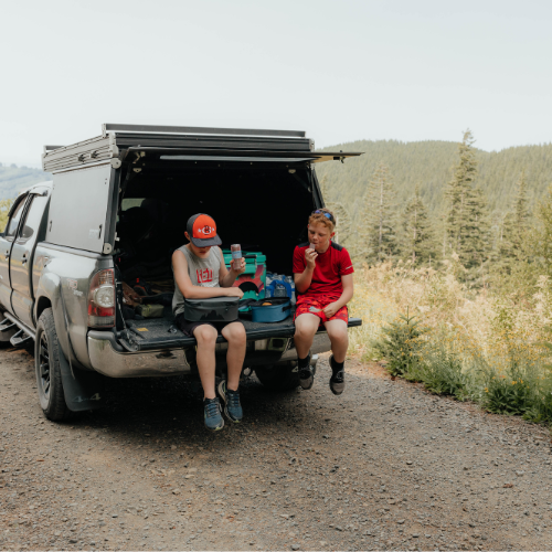 BACKCOUNTRY CLASS™ COOLER LUNCH BAG in turquoise blue with a dark top, carried at a truck tailgate picnic